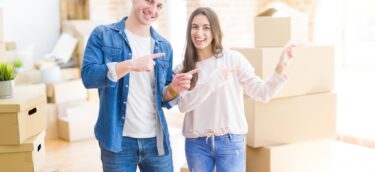 Beautiful young couple moving to a new house smiling and looking at the camera pointing with two hands and fingers to the side.